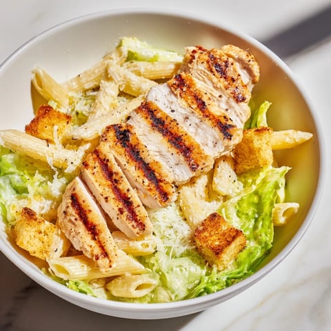 Tender grilled chicken slices rest atop a Caesar Pasta Chicken Bowl with al dente pasta, crisp romaine, and creamy dressing.