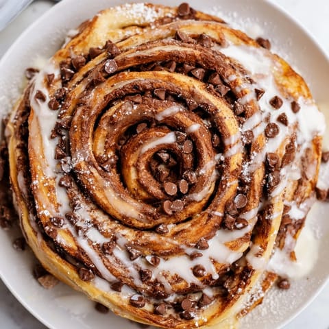 A close-up view reveals the soft, pillowy cinnamon swirl interior of a freshly baked Nutella Chocolate Chip Roll, with melted chocolate filling and a drizzle of glaze.