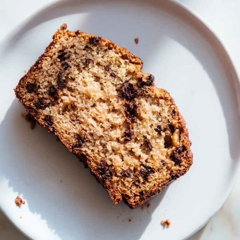 A slice of moist banana bread with chopped walnuts on a plate, beside a steaming mug of coffee for a cozy breakfast.  